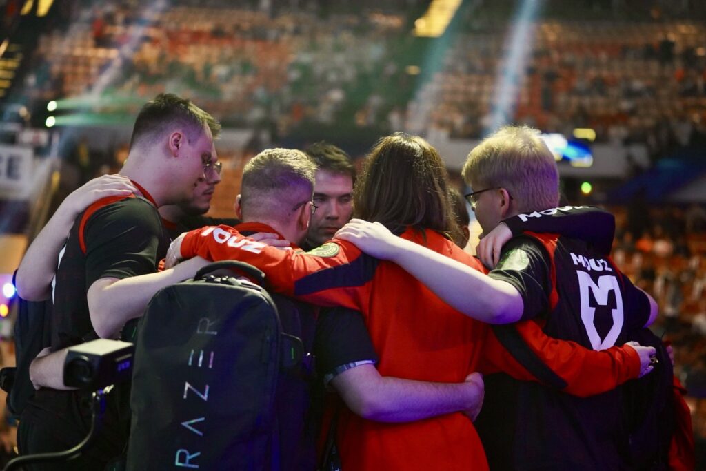 A team of professional Counter-Strike players embrace on stage at IEM Katowice 2024. You can see the stadium seats behind them.